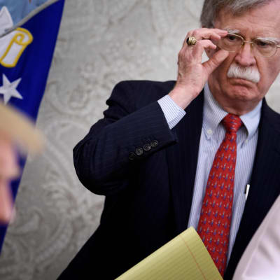 En kostymklädd man med maffig mustasch, John Bolton, rättar till sina glasögon och sneglar på Donald Trump som sitter framför honom.