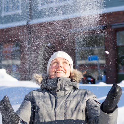 En vinterklädd kvinna ser glad ut och kastar upp snö i luften, snöhögar, trafikmärken och hus i bakgrunden.