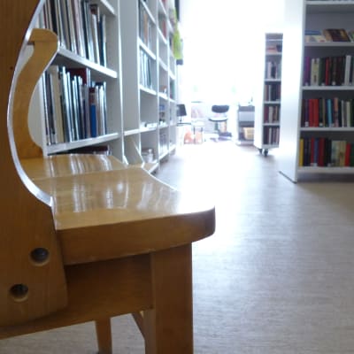 En gammal trästol i en ny ljus biblioteksmiljö. Vita hyllor fyllda med böcker. Ljus strömmar in genom ett fönster.