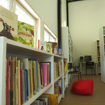 En hylla med barnböcker, andra hyllor med böcker i ett bibliotek, en röd säckdyna på golvet, i bakgrunden en dator på ett bord.