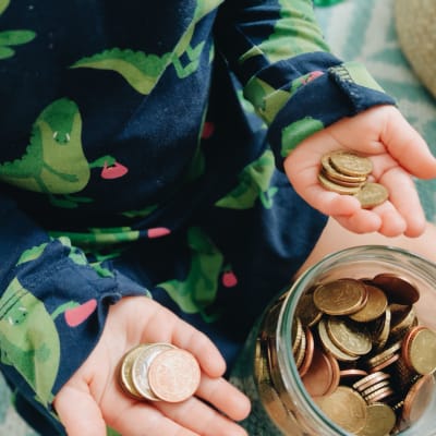 Ett barn håller mynt i båda händerna bredvid en glasburk full av mynt.