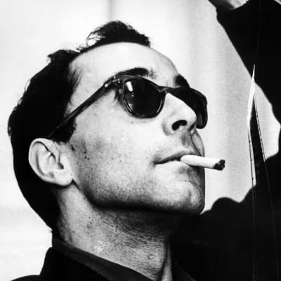 Mustavalkoinen kuva Jean-Luc Godardista aurinkolasit päässään tupakka suussa filminauhaa pidellen.