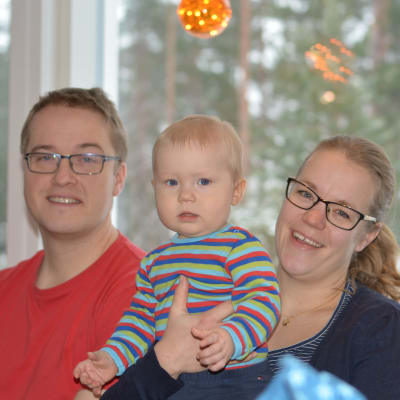 En familj sitter i soffa. En man och en kvinna och ett sju månader gammalt barn.Barnet är i mammans famn.  Alla ler.