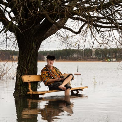 En äldre man sitter på en parkbänk som finns mitt i havet. Hans fötter är i vattnet.