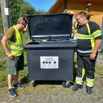 Två arbetare på en återvinningsstation tittar in i ett återvinningskärl för avlagda textilier