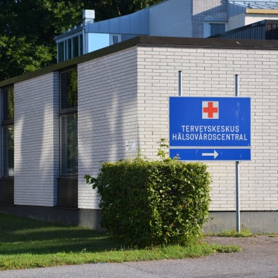 Hangö hälsocentral, en vit tegelbyggnad i 1-2 våningar. Soligt väder, grönt gräs. En blå skylt där det står Terveyskeskus Hälsovårdscentral.