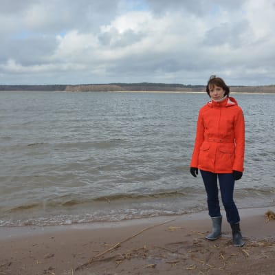 En kvinna i orange jacka och blå byxor står på en strand med vågor som rullar in. Hon heter Anna-Karin Almén.
