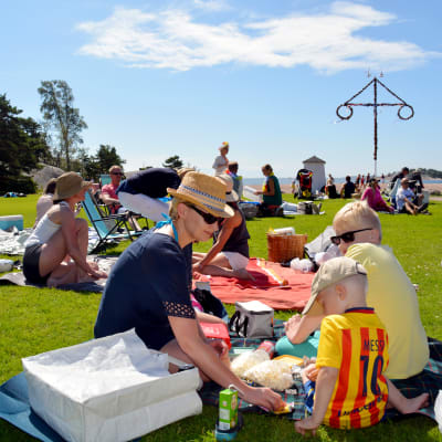 Människor sitter på gräset och har picknick. I bakgrunden skymtar en midsommarstång.