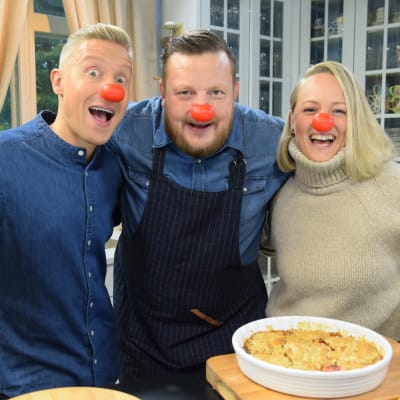 Två män och en kvinna med röda löstagbara näsor ler i ett kök med en äppelpaj framför sig.