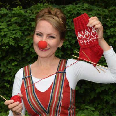 Kvinna med clownnäsa håller upp halvföärdigt stickad socka