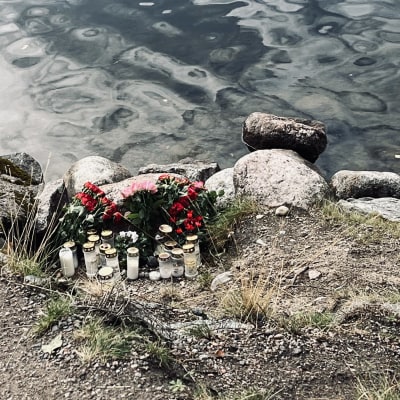 Hautakynttilöitä ja ruusukimppuja järven rantakivikossa.
