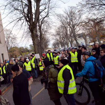 En stor grupp människor i gula reflexvästar står och demonstrerar framför en byggnad.