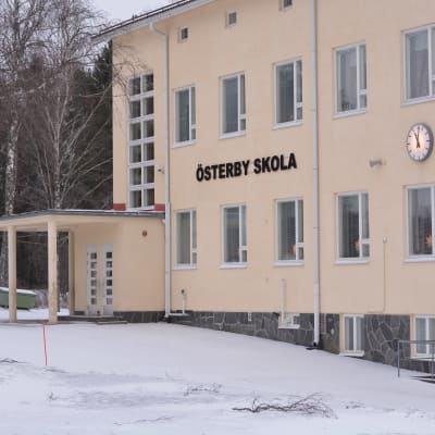 En skolbyggnad i flera våningar byggd i ljusmålad betong. Det står Österby skola på fasaden. På väggen finns också en stor, rund klocka. Vinter och snö, inga skolbarn.
