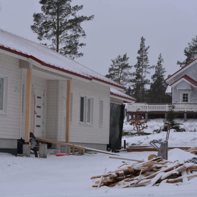 ett halvfärdigt hus, husbygge på gång. Vinter och snö. En hög med bräden. I bakgrunden ett färdigt, nybyggt hus.