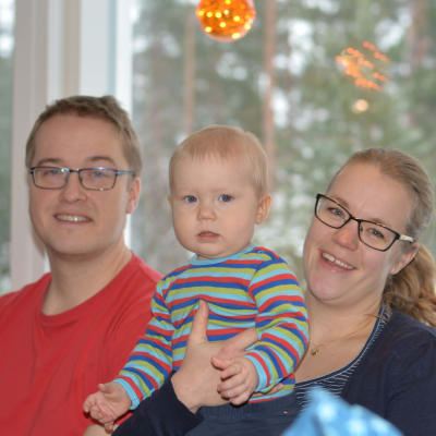 En familj sitter i soffa. En man och en kvinna och ett sju månader gammalt barn.Barnet är i mammans famn.  Alla ler.