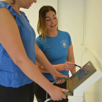 En kvinna  pekar med penna på en skärm och ler, en annan står med sidan till vid en apparat som mäter kroppens fett, muskler med mera.