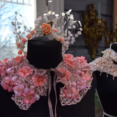 En dekorativ festkrage gjord av gammal, rosa, återvunnen spets och dekorerad med tygblommor och pärlor i olika rosa nyanser. Kragen kan användas till maskerad eller fest och är knuten runt halsen på en svart provdocka.