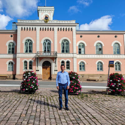 En man står på ett torg framför ett rosa stenhus.