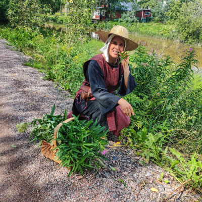 En kvinna sitter på huk bredvid en vägren och äter på en växt.
