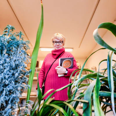 En kvinna i pink tröja står i ett rum med keramikskulpturer på hyllor. Hon  håller en bok i famnen. I förgrunden syns grönväxter.