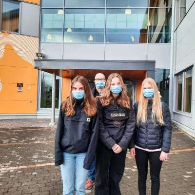Tre skolelever står framför ingången till Nickby hjärta. Bakom dem syns en äldre man, skolans rektor. Alla har munskydd på sig.