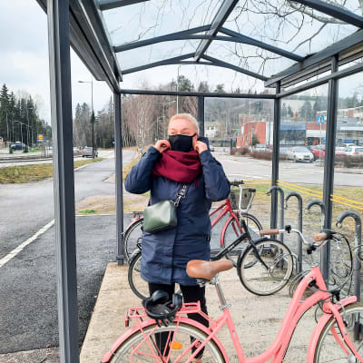 En kvinna sätter på sig ett munskydd. Hon står utomhus vid en cykelställning.