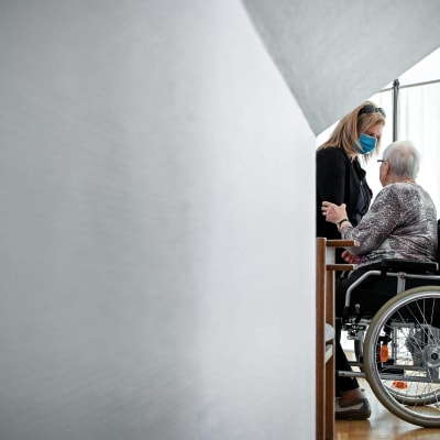 En person i munskydd hjälper en äldre person i rullstol.
