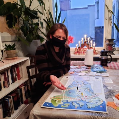 Kvinna i svart munskydd sitter vid köksbord och håller på med att kasta en tärning ovanpå en spelplan föreställande Sibbo.