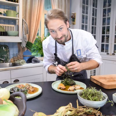 Man ser in i kameran i ett kök och dekorerar en matportion med örter.