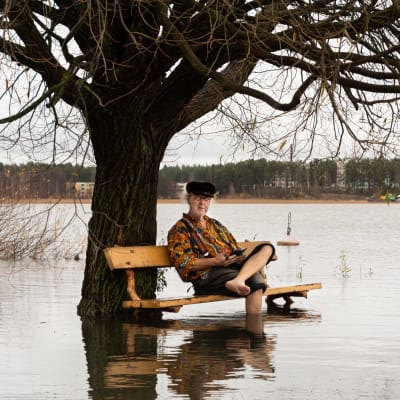 En äldre man sitter på en parkbänk som finns mitt i havet. Hans fötter är i vattnet.