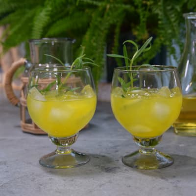 Två glas på fot med en knallgul dryck i. I glasen finns också isbitar och en kvist rosmarin.