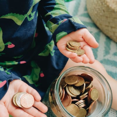 Ett barn håller mynt i båda händerna bredvid en glasburk full av mynt.
