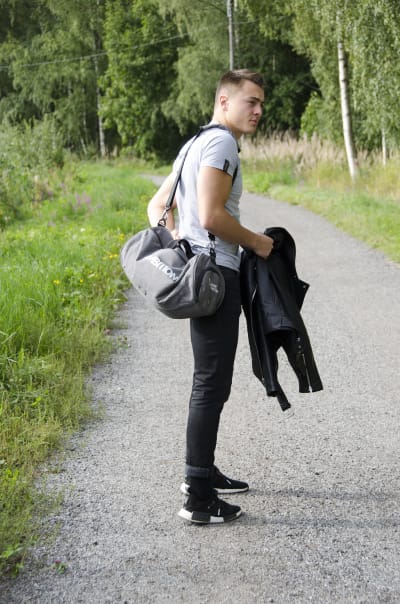 En man iklädd svarta jeans och en grå t-skjorta står på en grusväg kantad med grönt gräs och blommor. Han har ryggen snett mot betraktaren och tittar på något till sidan.