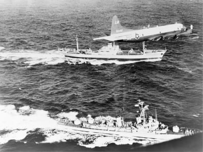 Ilmakuva merellä. Yhdysvaltain sota-alus seilaa neuvostoliittolaisen rahtialuksen vierellä. Ilmassa lentää yhdysvaltalainen lentokone.
