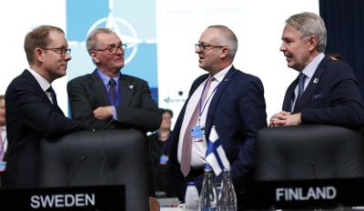 Fyra män i kostym diskuterar. I förgrunden Finlands flagga.