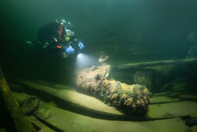 En dykare lyser med ficklampa ner på en gammal kanon i ett skeppsvrak.