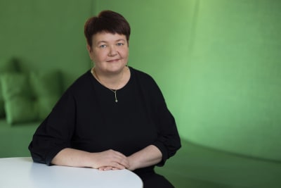 Nina Elomaa sitter iklädd en svart blus vid ett bord framför en enfärgat grön bakgrund, tittar in i kameran och ler.