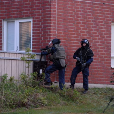 Poliser utanför ett rött tegelhus. De har tunga vapen och skyddsvästar på sig. 