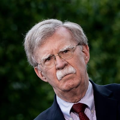 En man med grått hår, grå mustasch och glasögon, John Bolton, tittar åt sidan och ser allvarlig ut. 