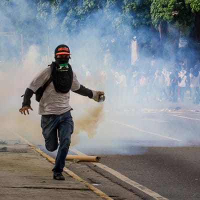 I förgrunden springer en demonstrant med ett rykande föremål i sin hand och skydd för munnen. I bakgrunden fler demonstranter.