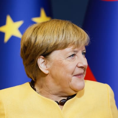En bild på Tysklands förbundskansler Angela Merkel. Hennes blick är riktad bort från kameran. I bakgrunden ser man EU:s flagga. 