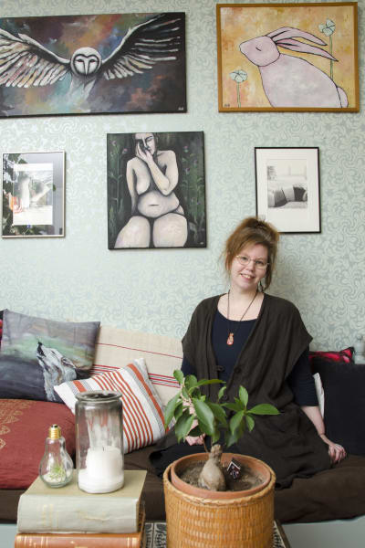 En kvinna som sitter på en soffa. Bakom hennes syns tavlor hon själv målat eller fotat. De föreställer djur och kvinnokroppen.
