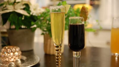 Två champagneglas med frosting av socker, ett med en ljusgul drink och ett med en mörkbrun drink. Den ena drinken är garnerad med en äppelskiva och den andra med en skiva ingefära.