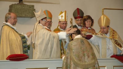 Blivande biskop Björn Vikström står på knä vid korskranket och många biskopar sträcker fram sina händer för att välsigna honom..