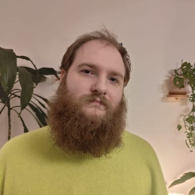 En man med skägg och grön skjorta med grönväxter i bakgrunden. 