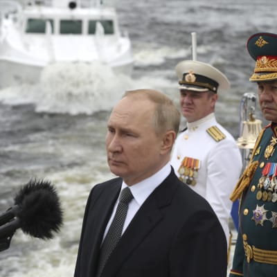 Vladimir Putin puhuu laivaston juhlatilaisuudessa. Taustalla vene ja koppapäisiä herroja.