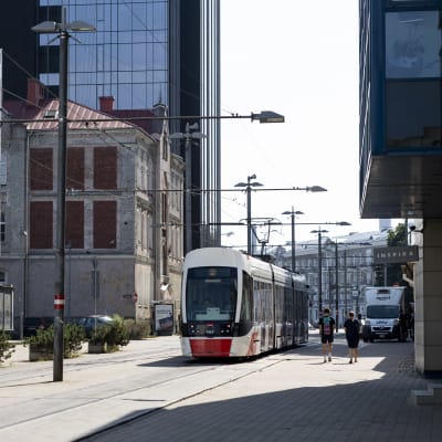 Punavalkoinen raitiovaunu kulkee kadulla, jonka varrella on sekä vanhoja tiilirakennuksia, että uusia lasipintaisia liikehuoneistoja. 