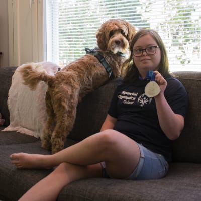 På bilden sitter Alina Skog med en logo-skjorta på sig och håller sin silvermedalj i handen. Bredvid henne på soffan sitter deras hund. 