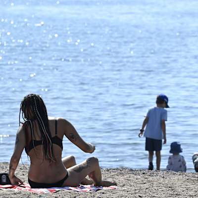 En kvinna med långt hår sitter på en filt på stranden. Hon har en svart bikini på sig. Vid havet sex barn som böjer sig ner och tittar nedåt.