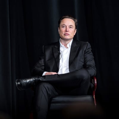 Elon Musk sitter på en stol med benen i kors. Han tittar framåt.